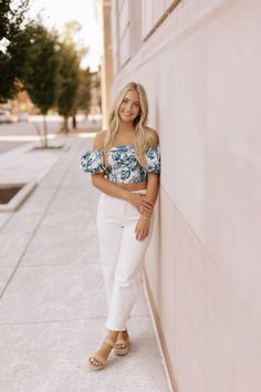 a woman leaning against a wall wearing white pants and a blue floral cropped top