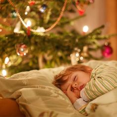 a small child is sleeping on a bed next to a christmas tree with lights in the background