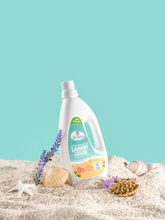 a bottle of laundry liquid sitting on top of sand next to shells and seaweed