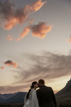 a bride and groom are kissing in front of the mountains at sunset with pink clouds