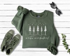 a green sweatshirt with trees on it next to some sneakers and sunglasses that says whatever is wonderful