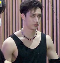 a young man wearing a black tank top and beaded necklace standing in front of a wooden wall