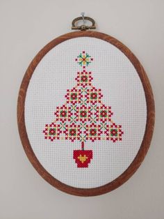 a cross stitch christmas tree hanging from a wooden hoop on a wall with an ornament in the shape of a heart