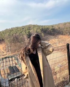 a woman standing in front of a fence wearing a trench coat and black shirt with her hand on her head