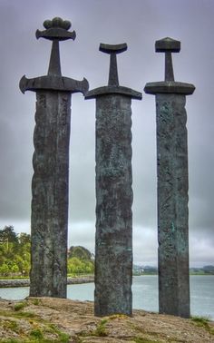 three stone pillars sitting on top of a grass covered hill next to the ocean in front of a cloudy sky