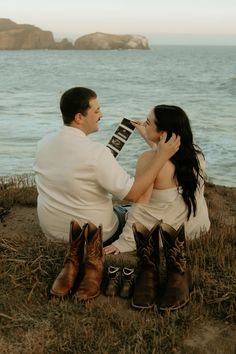 a man and woman sitting next to each other on the ground by the water with their feet up