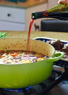 someone is pouring red wine into a green pot on the stove top, with meat and veggies in it