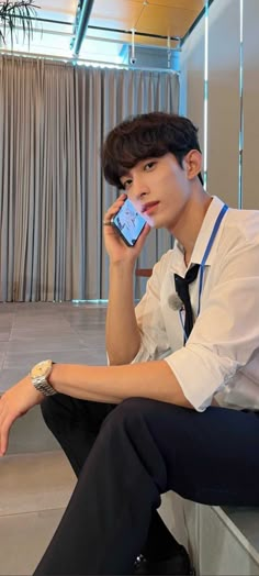 a young man sitting on the floor while holding a cell phone to his ear and looking at the camera