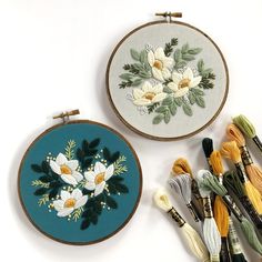 two embroidery hoops with flowers on them next to some thread and needle tips in the foreground