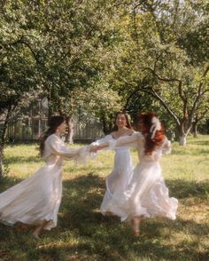 three women in long white dresses are playing with each other on the grass near trees