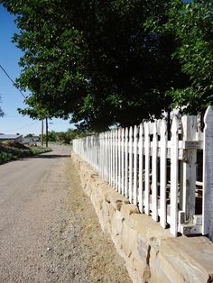 a white picket fence sitting on the side of a road next to a lush green tree