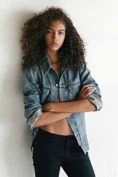 a woman with her arms crossed leaning against a wall and looking at the camera while wearing a jean jacket