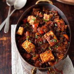 a pan filled with tofu and beans on top of a table next to spoons