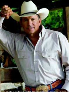 a man wearing a cowboy hat standing in front of a horse stable with his hand on his head