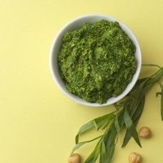 a white bowl filled with pesto next to green leaves and nuts on a yellow surface