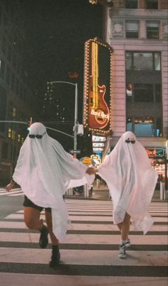 two people dressed in ghost costumes crossing the street at night with neon signs behind them