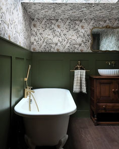 a bathtub and sink in a green bathroom with wallpapered walls, wood flooring and wooden cabinet