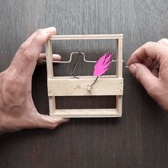 two hands holding a small wooden box with a pink feather in it on a table