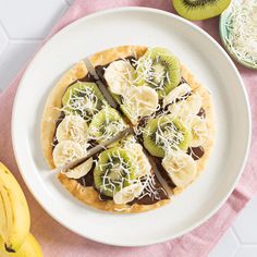 a white plate topped with a pizza covered in kiwis and cheese next to sliced bananas