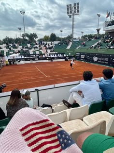 people sitting in chairs watching a tennis match on a court with an american flag blanket