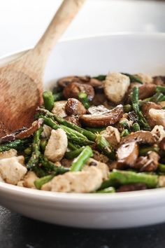 a white bowl filled with asparagus, mushrooms and other vegetables next to a wooden spoon