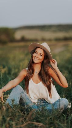 a woman sitting in tall grass wearing a hat and smiling at the camera with her hands behind her head