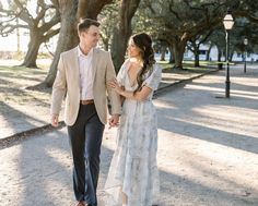 a man and woman holding hands walking through the park in front of some large trees