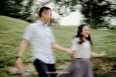 a man and woman holding hands while walking in the grass near a stone wall with trees behind them