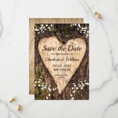 save the date card with wooden heart surrounded by greenery and tiny white flowers on a marble background