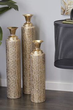 three gold vases sitting next to each other on a wooden floor near a potted plant