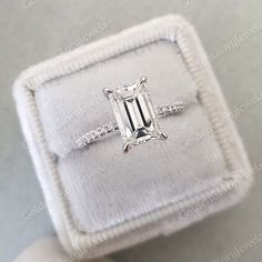 an emerald cut diamond ring in a white velvet box with diamonds around the band and side stones