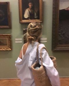 a woman in a white dress is carrying a wicker basket and looking at paintings on the wall