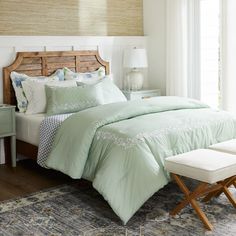 a bed with green comforter and pillows in a bedroom next to a window on top of a rug