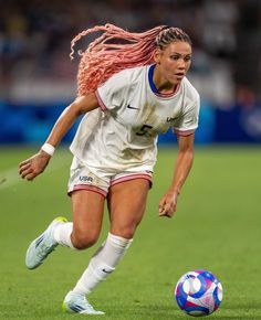 a female soccer player is running with the ball in her hand and pink dreadlocks on her hair