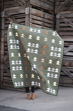 a woman holding up a quilt with trees on it in front of a wooden building