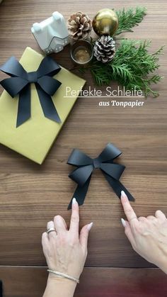 two hands holding black paper bows on top of a wooden table next to presents and pine cones