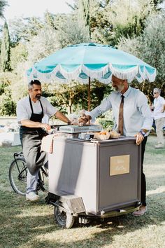 two men in aprons are cooking food on an outdoor grill with umbrella over it