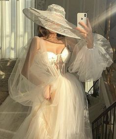 a woman in a white dress and hat taking a selfie with her cell phone