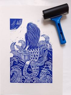 a blue and white ink drawing of a mermaid on paper with a rubber stamper next to it