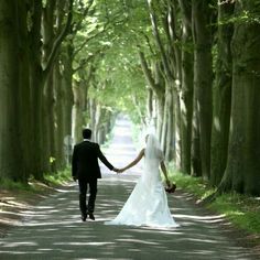 a bride and groom holding hands while walking down a tree lined road in the woods