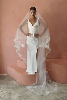 a woman wearing a white wedding dress with a veil on her head standing in front of a pink wall