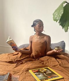 a woman sitting on a bed reading a book and drinking coffee while holding a cup