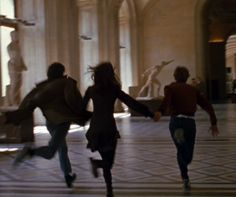 three people are running in the middle of a room with statues on either side of them