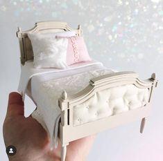 a hand holding a miniature white bed with pink pillows