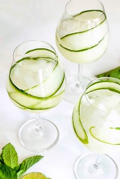 three glasses filled with cucumber and lemonade on top of a white table