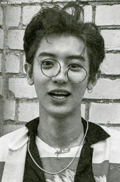 a young man wearing glasses standing in front of a brick wall