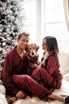 a man and woman in plaid pajamas sitting next to a christmas tree with their dog