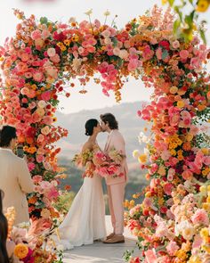 two brides kissing in front of a floral arch with pink and yellow flowers on it