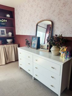 a dresser with a mirror and pictures on it in front of a wallpapered room