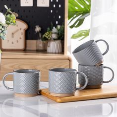 three grey coffee mugs sitting on top of a wooden cutting board next to a vase with flowers